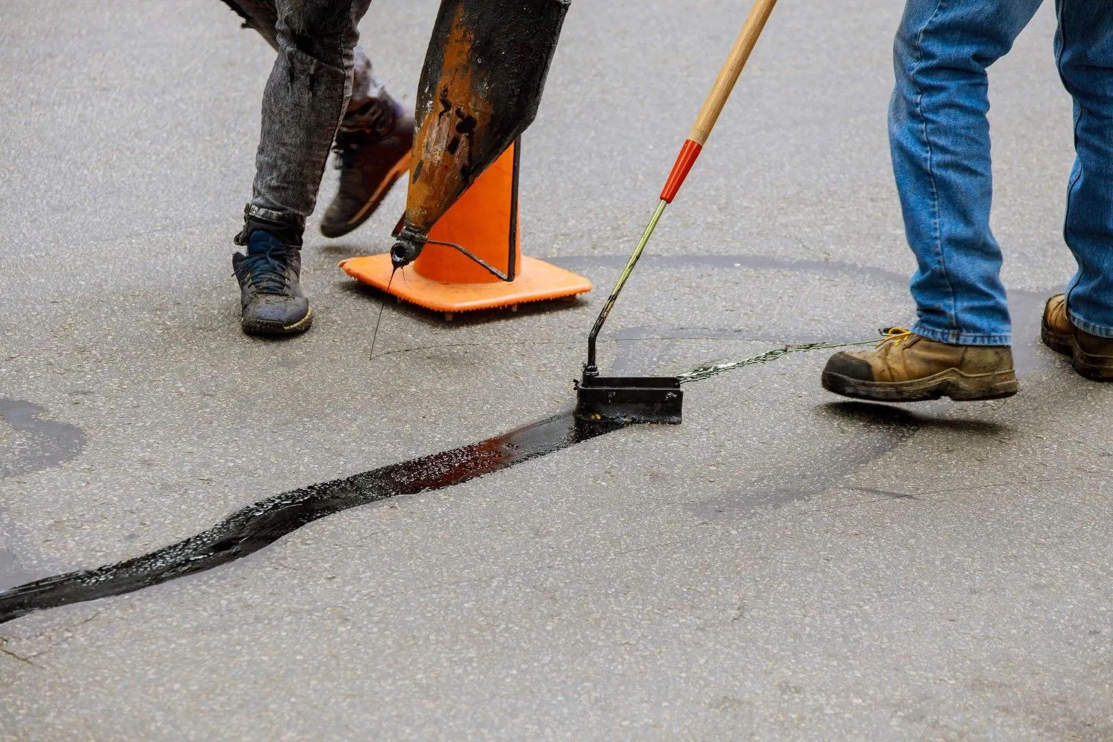 Workers sealing a long crack in a concrete driveway using a squeegee and hot sealant